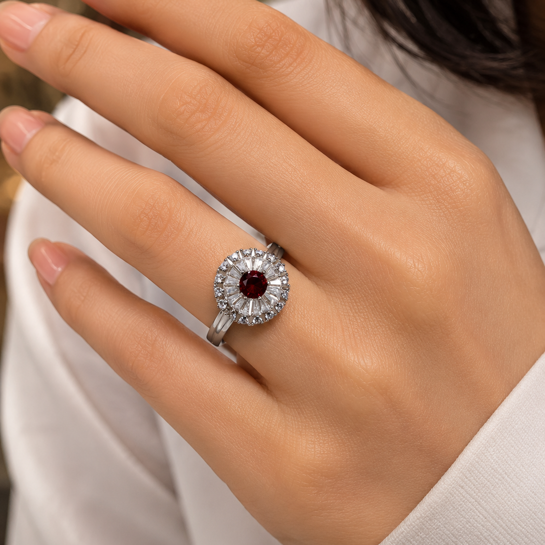 A close-up of a hand wearing a silver-toned ring with a round-cut dark red ruby center stone surrounded by a halo of small clear stones.