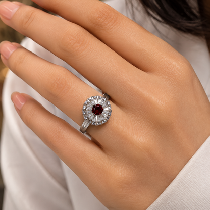 A close-up of a hand wearing a silver-toned ring with a round-cut dark red ruby center stone surrounded by a halo of small clear stones.