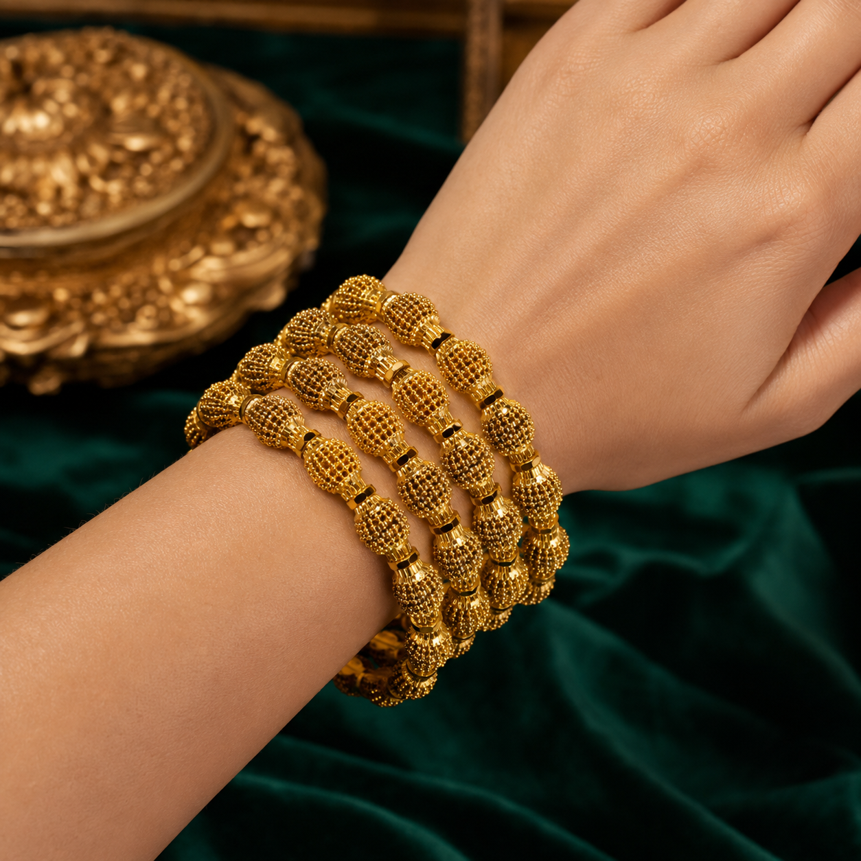A close-up of a person's wrist wearing four stacked gold beaded bracelets with textured, knotted details, set against a dark green velvet background with an ornate gold decorative object in the upper left.