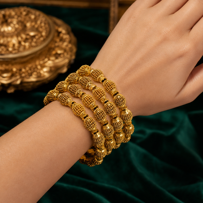 A close-up of a person's wrist wearing four stacked gold beaded bracelets with textured, knotted details, set against a dark green velvet background with an ornate gold decorative object in the upper left.