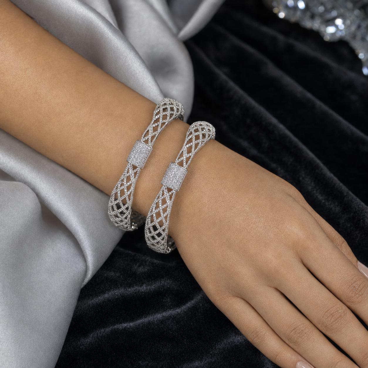 Two silver-toned filigree bangle bracelets with pavé-set clear stones, shown worn on a model's wrist against a backdrop of silver satin and black velvet.