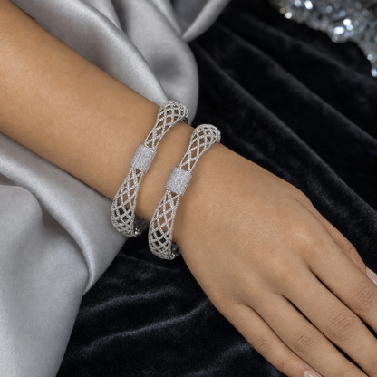 Two silver-toned filigree bangle bracelets with pavé-set clear stones, shown worn on a model's wrist against a backdrop of silver satin and black velvet.