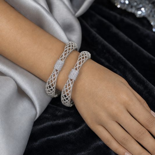 Two silver-toned filigree bangle bracelets with pavé-set clear stones, shown worn on a model's wrist against a backdrop of silver satin and black velvet.