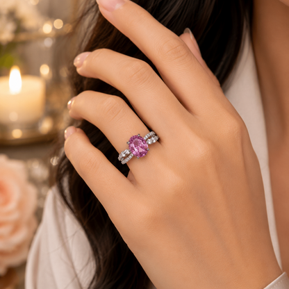 A close-up lifestyle shot of a woman's hand wearing a silver ring with a prominent oval purple gemstone and small side stones, set against a warm, softly blurred background with a lit candle.