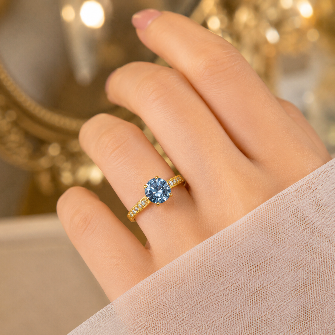 A close-up of a hand wearing a yellow gold ring with a round-cut light blue gemstone and small side stones, set against a softly blurred warm-toned background.
