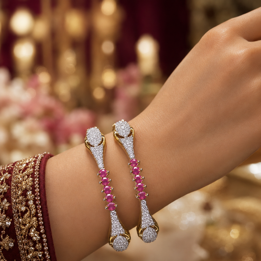 A close-up of a person's wrist wearing two gold-toned bangles adorned with pavé white stones and a row of pink gemstones, set against a softly blurred warm background.