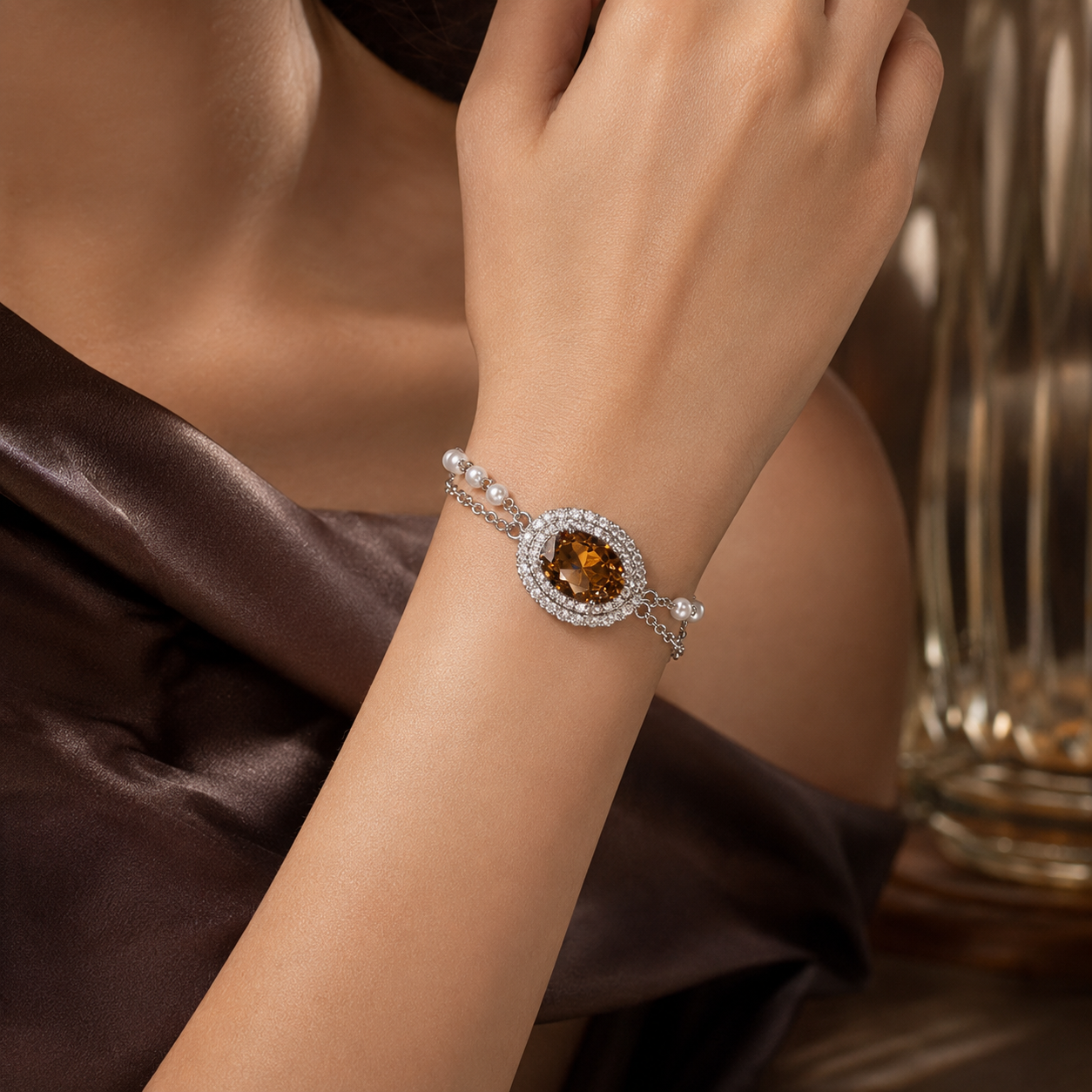 A close-up lifestyle shot of a woman's wrist wearing a delicate silver bracelet. The bracelet features a central oval brown gemstone surrounded by a halo of small clear stones, with a side detail of white pearls, set against a dark brown satin dress and a blurred glass background.