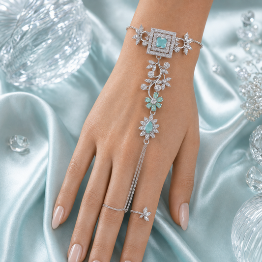 A silver-toned chain hand bracelet with floral motifs and light blue-green stones, shown worn on a hand over a shiny pale blue satin fabric with decorative crystal-like props.
