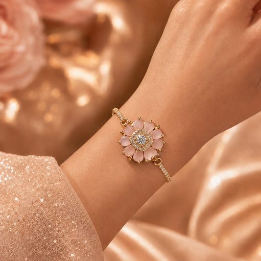 A delicate gold-tone bracelet with a lotus flower charm featuring pale pink petals and a clear central stone, shown worn on a wrist against a soft rose-gold satin background.