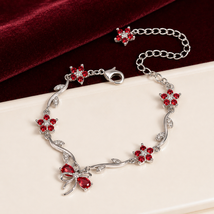 A silver-toned vine bracelet decorated with red crystal flowers and a bow-shaped centerpiece, displayed on a cream surface with a dark red velvet cloth in the background.