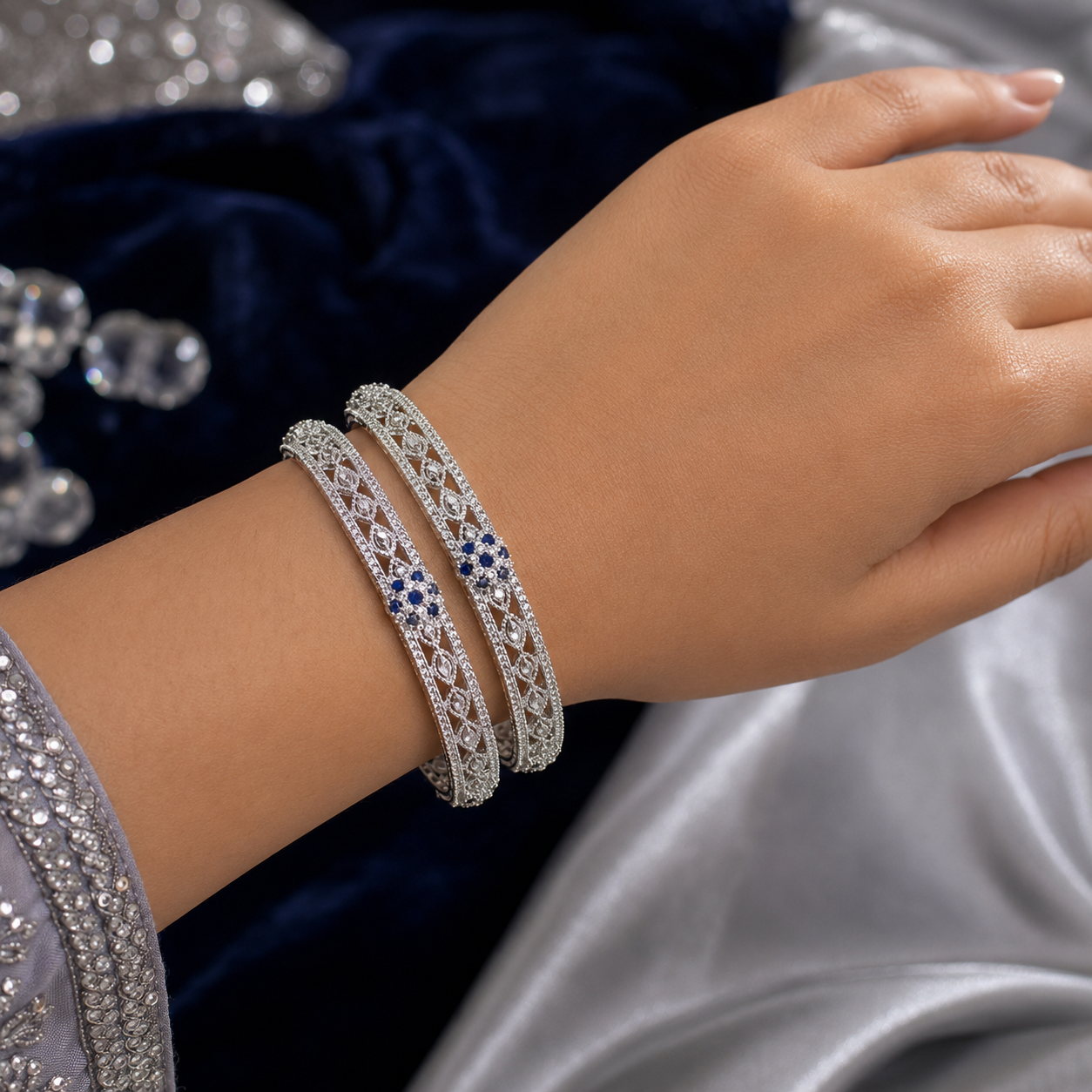 A close-up of a person's wrist wearing two silver-toned bangle bracelets adorned with pavé-set clear stones and a small blue gemstone accent, posed against a dark blue velvet and silver satin background.
