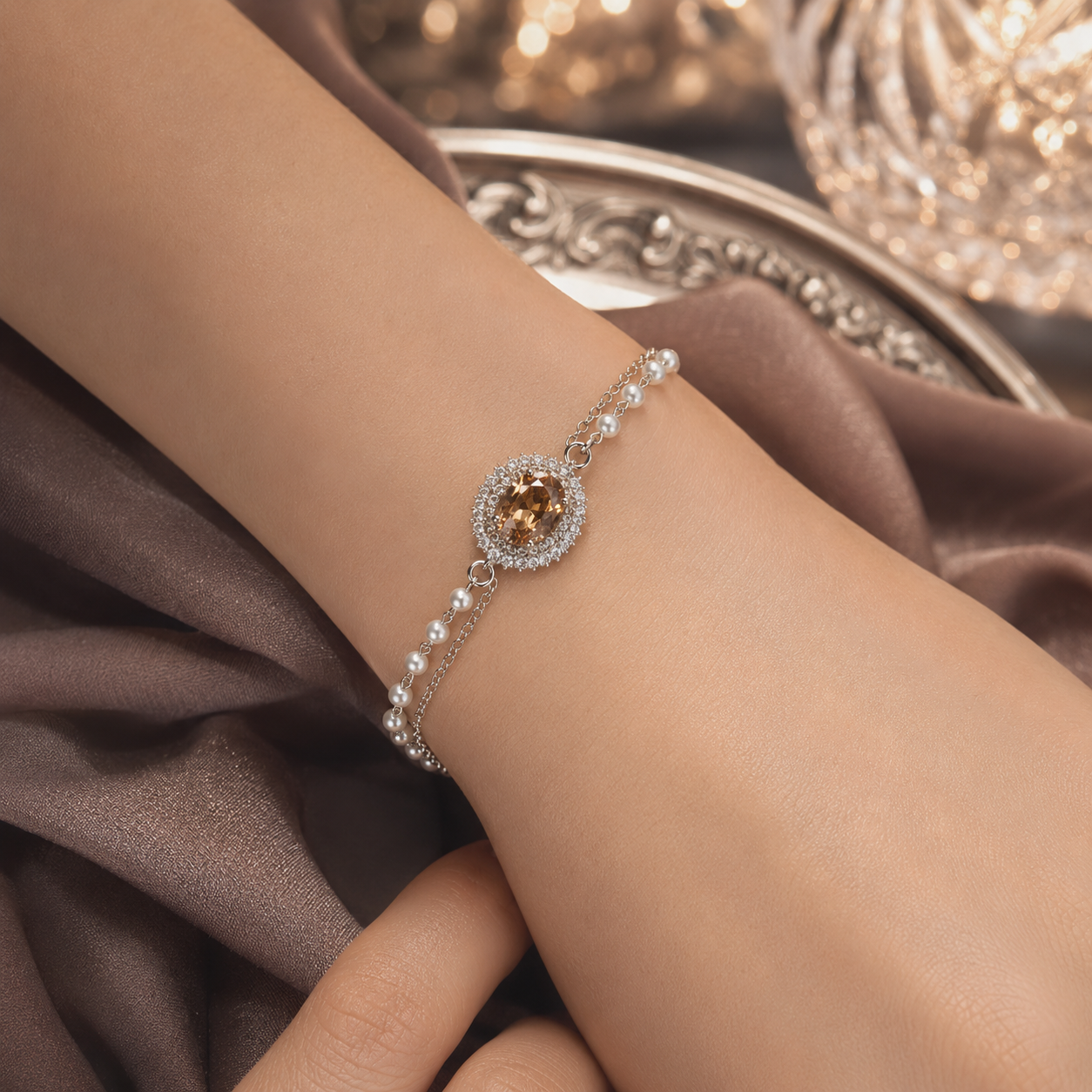 A delicate silver-toned bracelet with a central round amber-brown gemstone surrounded by small clear stones, shown worn on a person's wrist against brown draped fabric.